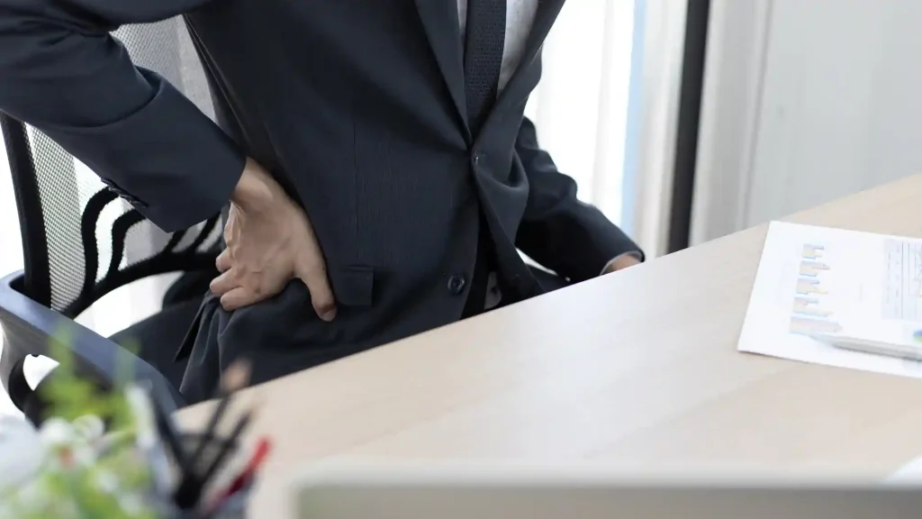 Person in a suit holding their lower back in pain while sitting at a desk